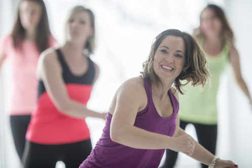 Group of women are taking an aerobic dance fitness class together at the gym. One woman is smiling and looking at the camera.